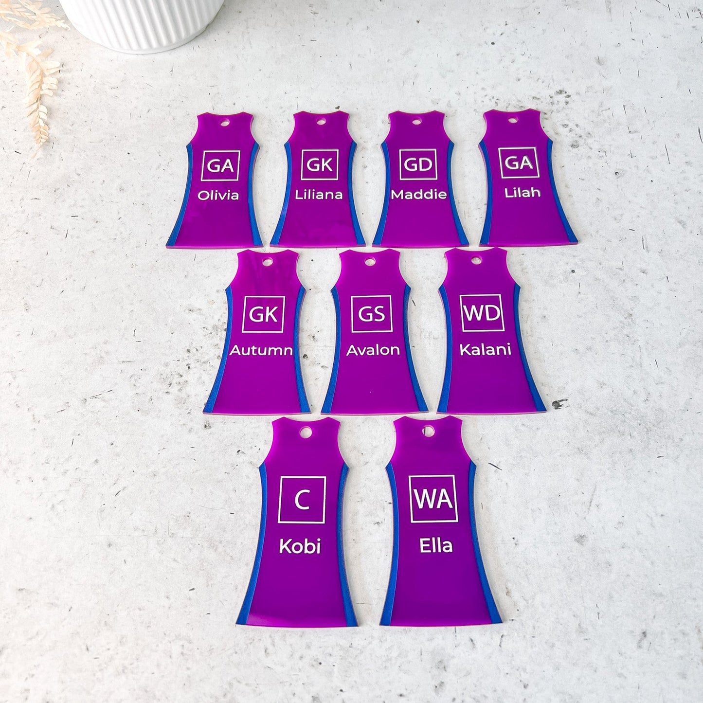 A collection of purple acrylic keyrings shaped like netball dresses, each with different names and initials engraved and painted on them, displayed against a light background.