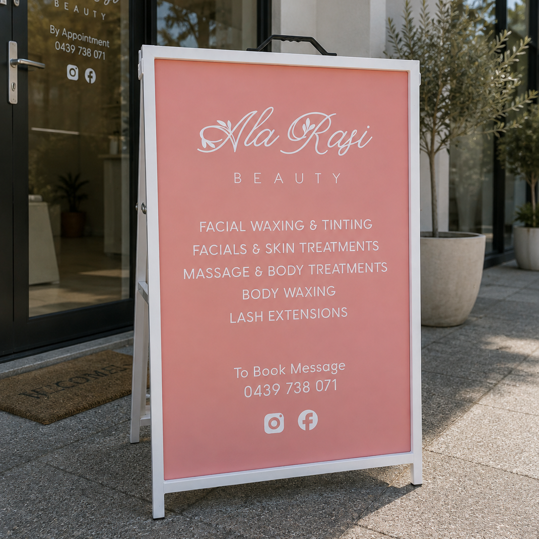 A pink A-frame sidewalk sign with white lettering for Ala Rasi Beauty, placed outside the salon on a paved walkway.